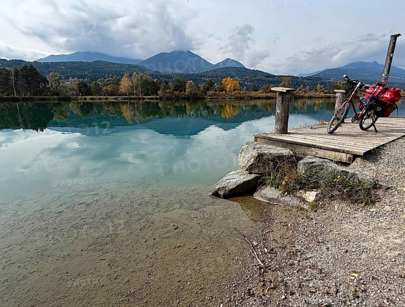 A bicycle is leaning on a footbridge over a still lake, mountains and clouds are reflected in the clear water, Drau, Drau cycle path, Carinthia, near Villach, Austria