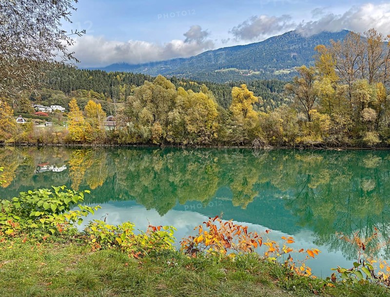 Calm river reflecting autumnal trees and mountains under cloudy sky, Drau, Drauradweg, Carinthia, near Villach, Austria