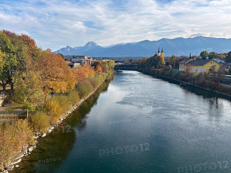 River through a city, autumn trees and mountains in the background under a clear sky, Drau, Drau cycle path, Carinthia, near Villach, Austria