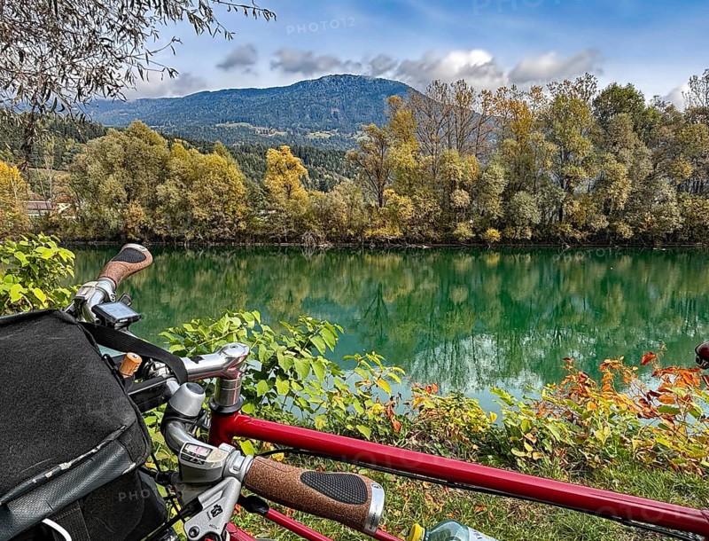 View over the handlebar of a shimmering green river in front of autumn trees and mountains, bicycle tourism, Drau, Drau cycle path, Carinthia, near Villach, Austria