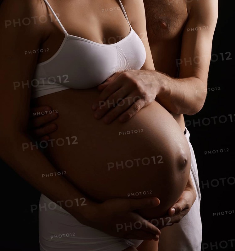Pregnant young long haired woman staying close to her husband both tenderly looking at her belly and holding their hands on it