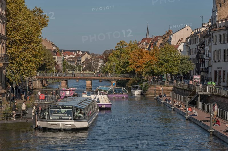 Tourist boat docks on the Ill and transports visitors who enjoy the view of Strasbourg's sights and trees in autumn. The scenery is peaceful and sunny. Bas Rhin, Alsace, France