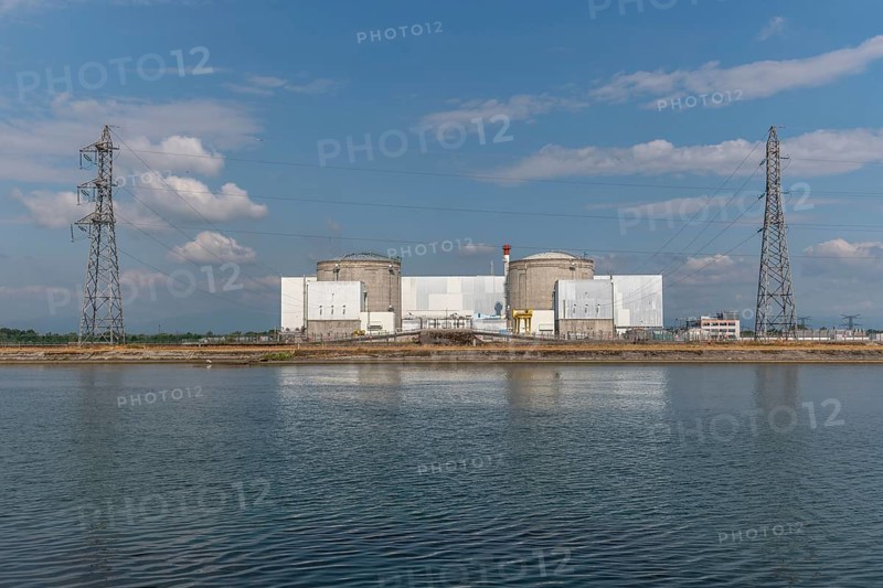 A large industrial nuclear power plant is located on a quiet water front. Two tall cylindrical structures dominate the skyline, with overhanging power lines and distant clouds that ennoble the blue sky. Fessenheim, Haut Rhin, Alsace, France
