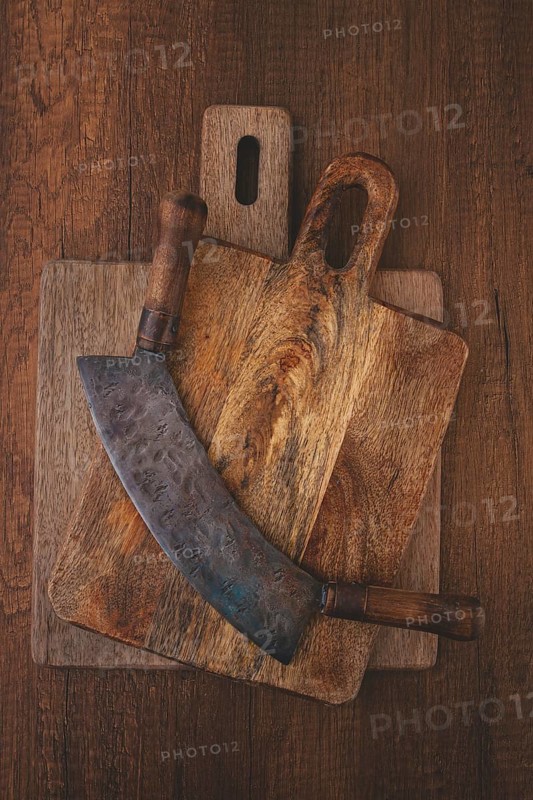 A large curved knife rests on top of two wooden cutting boards on a rustic surface. The knife shows wear, while the boards display unique grain patterns, adding character to the scene