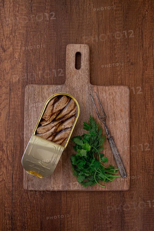 An open can of sprats, appetizer, on a wooden cutting board, with a fork and herbs, top view