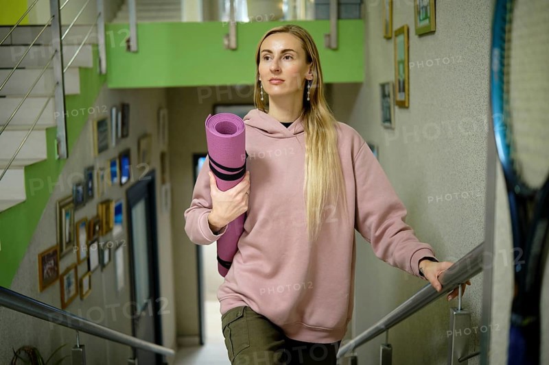 A woman with long hair is walking up the stairs of a vibrant yoga studio. She holds a rolled yoga mat and wears a casual pink sweatshirt, ready for her yoga practice
