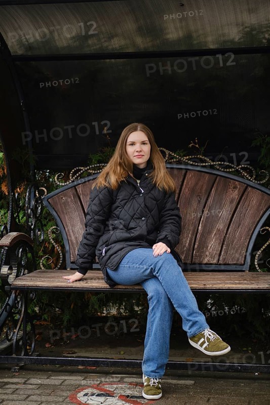 A young woman sits calmly on a wooden bench in a park surrounded by autumn foliage. She wears a black jacket and blue jeans, enjoying the peaceful atmosphere in a serene setting
