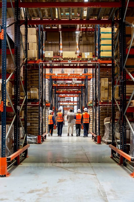 Workers in hard hats and reflective vests walk down a high rack warehouse aisle filled with stacked boxes and pallets, illustrating teamwork, safety and efficient supply chain operations