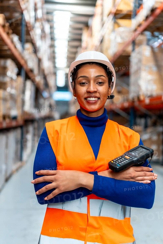 African american woman worker wearing a safety vest and hard hat, holding a portable scanner, standing proudly with arms crossed in a large modern logistics warehouse