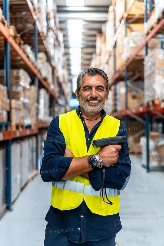 Experienced warehouse professional in a high visibility vest, smiling confidently with folded arms and barcode scanner in a neat logistics distribution aisle, ready for inventory operations