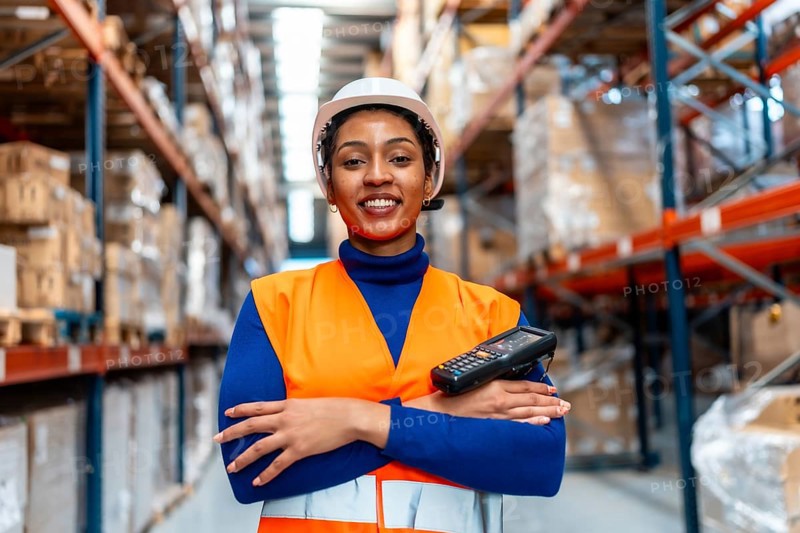 Female logistics operator wearing safety vest and hard hat, standing confidently with a barcode scanner in a modern distribution warehouse full of racks and inventory