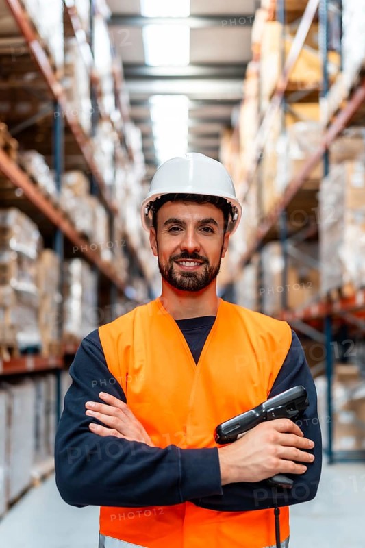 Warehouse worker in safety vest and hard hat smiles at camera while holding a barcode scanner in a modern logistics facility, conveying efficient inventory control and fulfillment operations