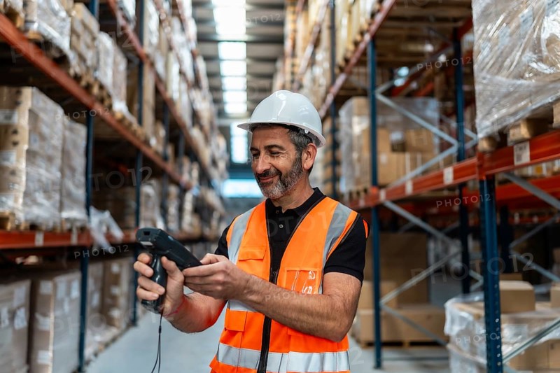 Male logistics worker wearing a hard hat and safety vest, scanning packages on shelves in a large distribution warehouse, managing stock and supply chain operations