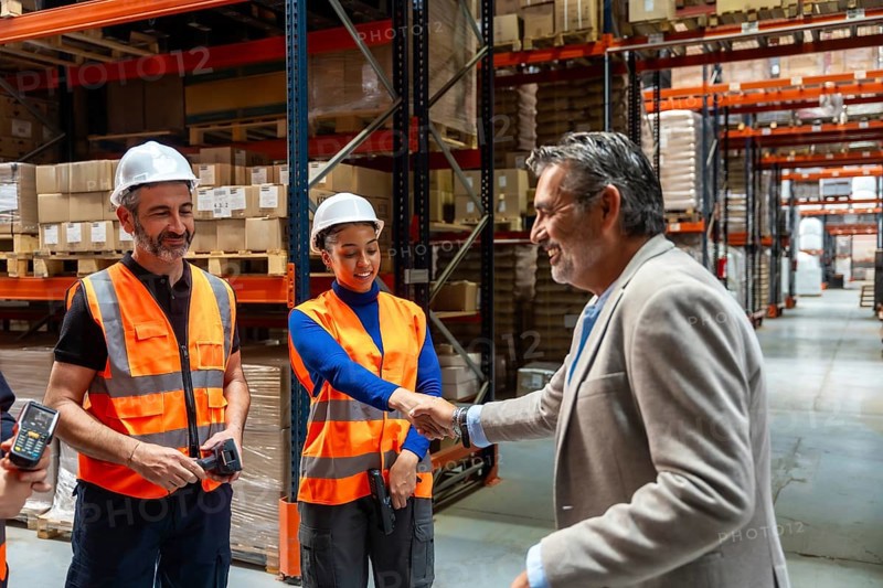 Manager shaking hands with a woman warehouse worker while male team members smile, symbolizing partnership, collaboration, and successful business agreement in a logistics facility