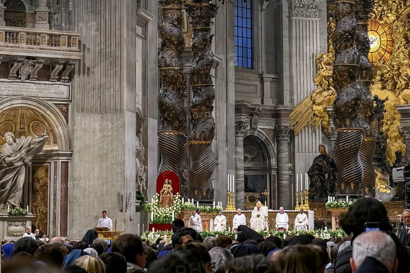 View of heads of faithful in the foreground at the bottom of the picture Pope Saint Father with white robe and mitre on head between columns of canopy by Bernini over the tomb of Peter celebrates Saint Mass in St. Peter's Basilica, Basilica of St. Peter, Vatican, Rome, Lazio, Italy