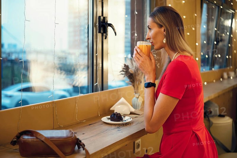 A woman in a red dress sits by the window of a cafe, sipping a warm drink and enjoying a dessert. Warm lights create a relaxed vibe as she gazes out at the bustling city during the evening