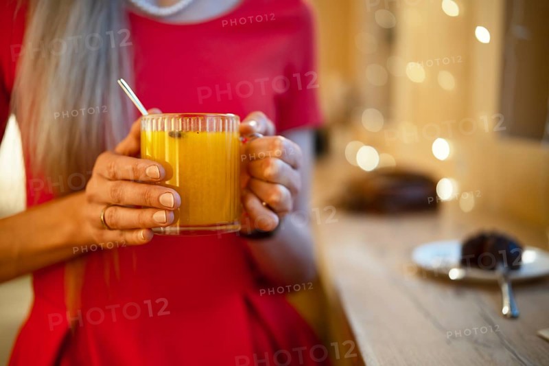 A woman in a bright red dress holds a glass of warm orange drink, enjoying a relaxing moment. Soft lights create a warm atmosphere. In the background, a dessert plate is visible on the table