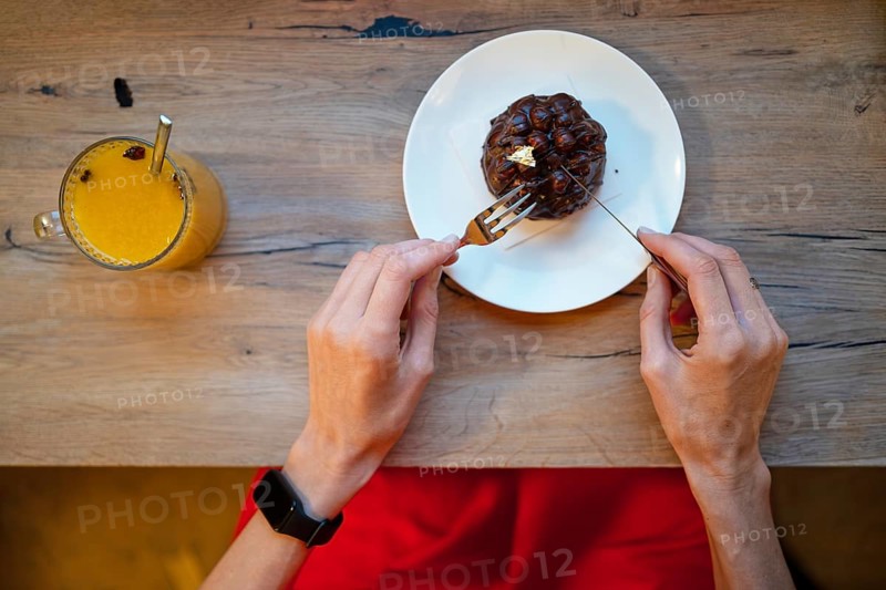 A person sits at a wooden table, ready to enjoy a delicious dessert. A fork is poised over the sweet treat, while a colorful drink sits nearby, creating a perfect evening indulgence
