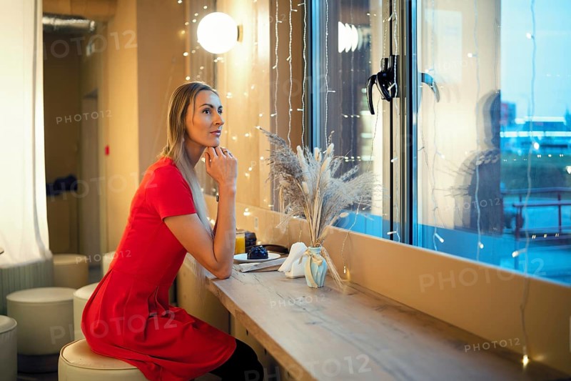 A woman dressed in a red outfit sits by a window in coffee shop, deep in thought. Fairy lights illuminate the space as she enjoys a quiet moment in a cafe. The atmosphere is peaceful and reflective