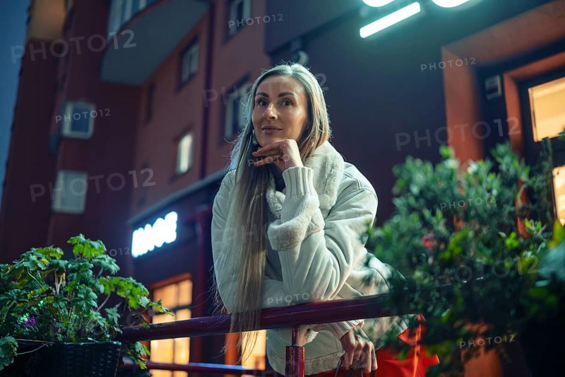 A woman stands thoughtfully on a balcony railing, dressed warmly for the evening chill. The city lights glow softly in the background as she gazes off into the distance, surrounded by greenery