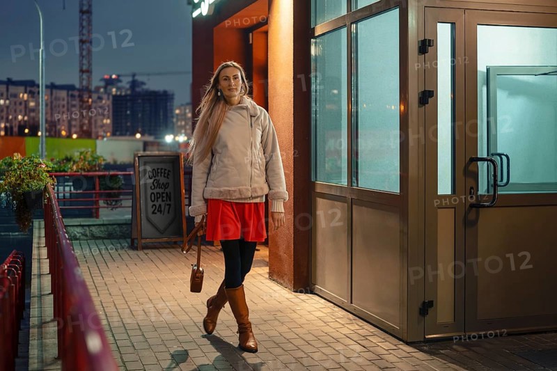 A woman strolls outside a coffee shop in the evening. She is dressed in a bright red dress, brown boots, and a cozy jacket