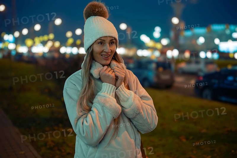 A woman stands outside on a city street during the evening, wearing a warm jacket and a hat with a pompom. She smiles while surrounded by twinkling street lights and distant cars