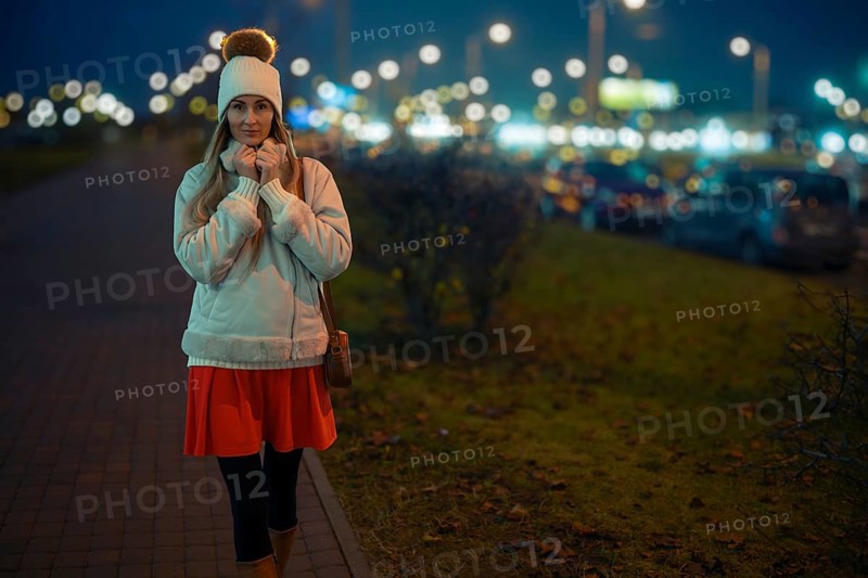 A woman walks on a city sidewalk at night, wearing a warm white jacket and a beanie. The street is filled with bright lights and parked cars, creating a lively urban atmosphere