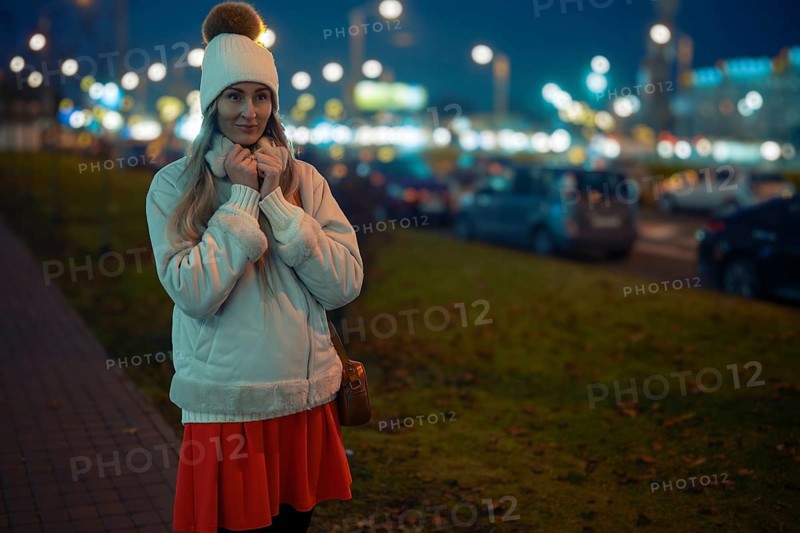 A woman stands outside on a cool evening, wearing a warm jacket and beanie. She smiles softly while holding her hands close to her face, illuminated by bright city lights