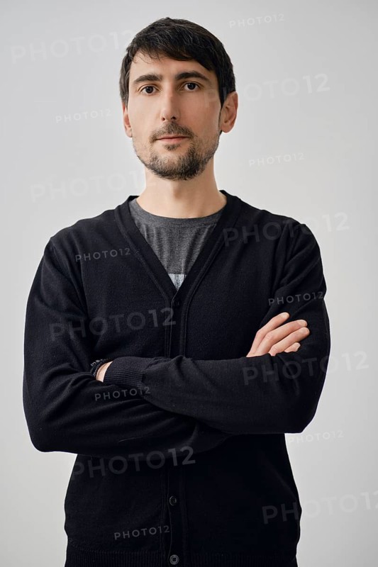 A man stands with arms crossed, portraying confidence. He wears a black cardigan over a gray shirt. The background is simple and light, emphasizing his serious expression during the day