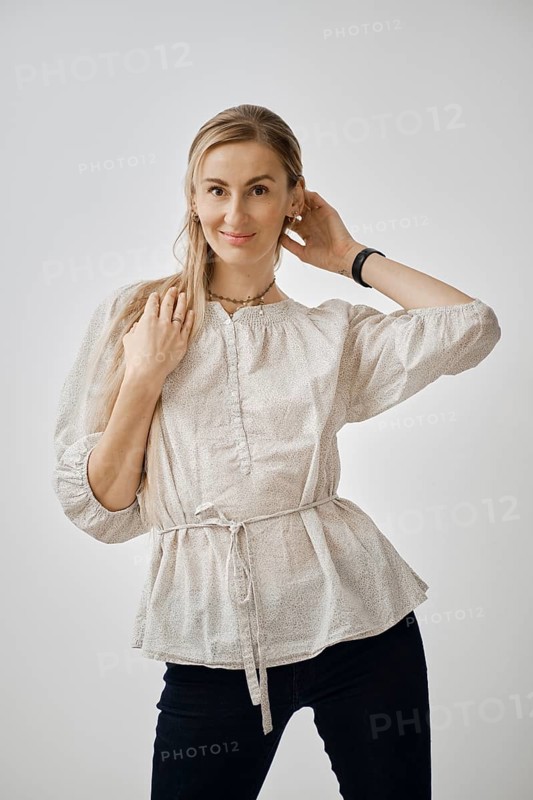A playful woman stands in a bright studio, wearing a fashionable light blouse and dark pants. She smiles while playing with her hair, showcasing a relaxed and cheerful mood