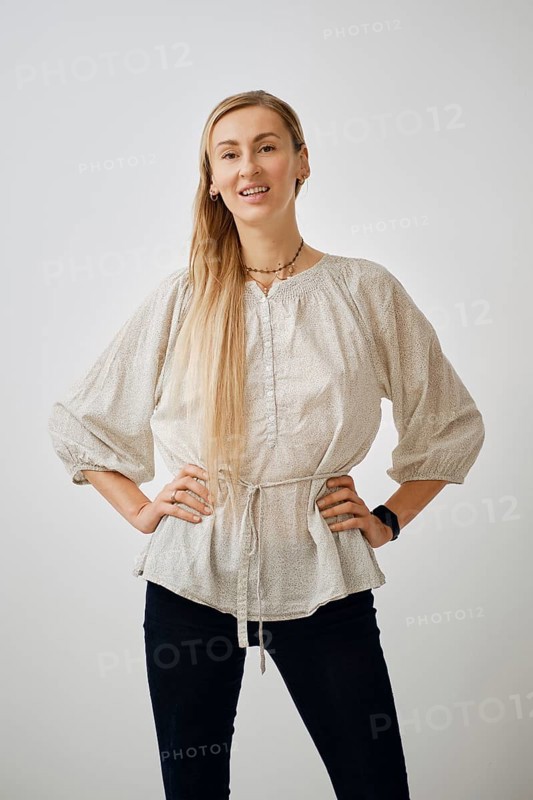 A woman stands confidently in a plain studio, wearing a light, textured blouse and dark pants. She smiles at the camera, showcasing a friendly and approachable demeanor