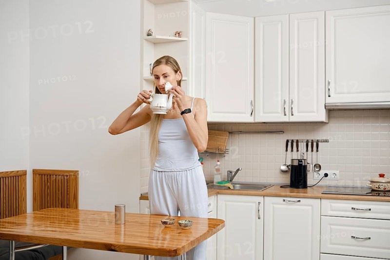 In a modern kitchen, a young woman in light clothing inhales the aroma from freshly brewed tea from a teapot. Soft morning light fills the room as she stands by a wooden table