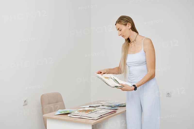 A woman with long hair stands in a bright, minimal room, looking through art sketches. She focuses on the details of the drawings while surrounded by various creative works on the table
