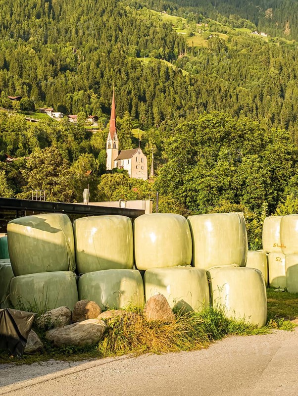 Agricultural structures with hay bales sealed in plastic near a farm on the outskirts of Oetz in Ötztal, Tyrol, Austria, with the parish church in the background