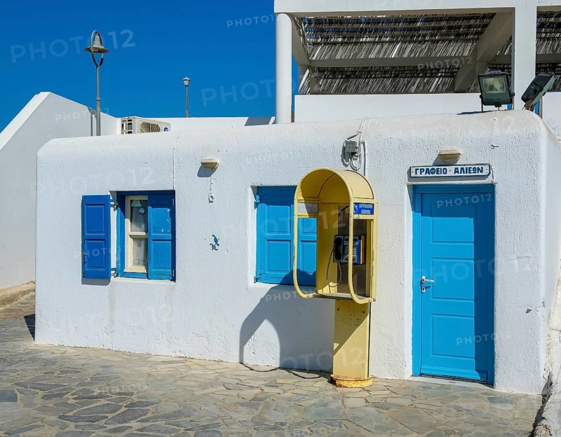 Mykonos, Cyclades, Greece - Old yellow telephone box in front of white house with blue shutters in the old town alleys of Mykonos Town. Mykonos is part of the Cyclades archipelago in the Aegean Sea