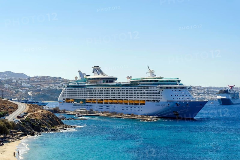 Mykonos, Cyclades, Greece - Voyager of the Seas cruise ship is located in Mykonos Harbour Bay. In front, tourists bathe on the sandy beach of Paralia Agios Stefanos. In the back is the old town of Mykonos