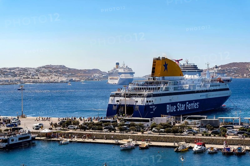 Mykonos, Cyclades, Greece - Mykonos New Port, a Blue Star Ferries ferry, is located on the quay in the new port in front of the skyline of Mykonos Town. In front, a taxi boat a sea bus takes tourists to the old town. At the back, there are more cruise ships in the bay in front of Mykonos City with a view of the old town with the windmills. Cruise ships bring tourists to the old town