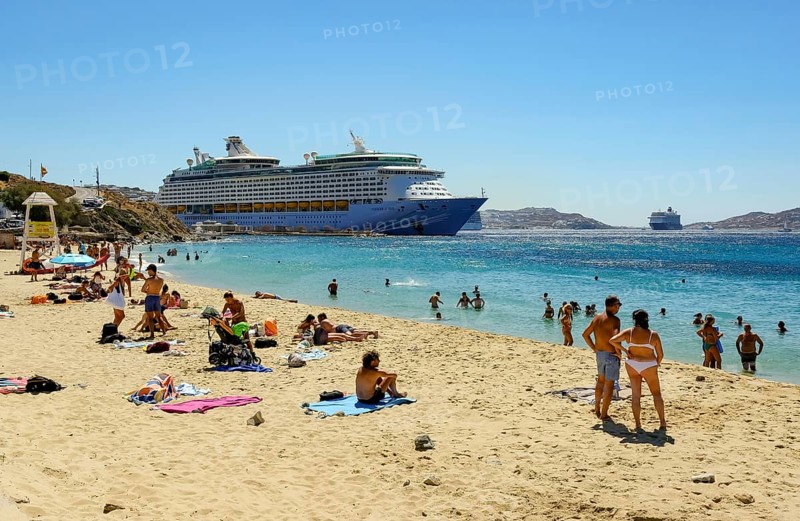Mykonos, Cyclades, Greece - Voyager of the Seas cruise ship is located in Mykonos Harbour Bay. In front, tourists and locals bathe on the sandy beach of Paralia Agios Stefanos