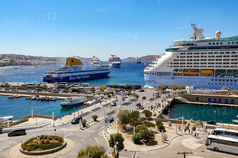 Mykonos, Cyclades, Greece - Mykonos New Port, a Blue Star Ferries ferry and a cruise ship are located on the quay in the new port in front of the skyline of Mykonos Town. At the back, there are more cruise ships in the bay off Mykonos City with a view of the old town with the windmills. Cruise ships bring tourists to the old town