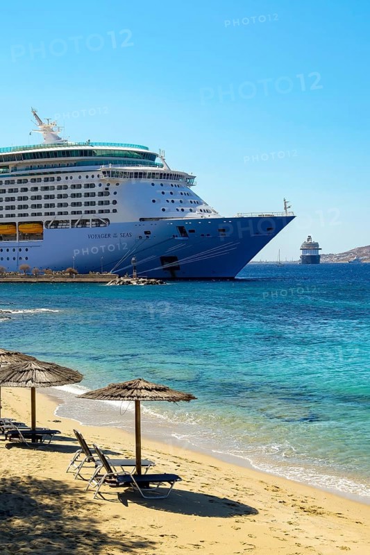 Mykonos, Cyclades, Greece - Voyager of the Seas cruise ship is located in Mykonos Harbour Bay. Front beach chairs and umbrellas on the sandy beach of Paralia Agios Stefanos