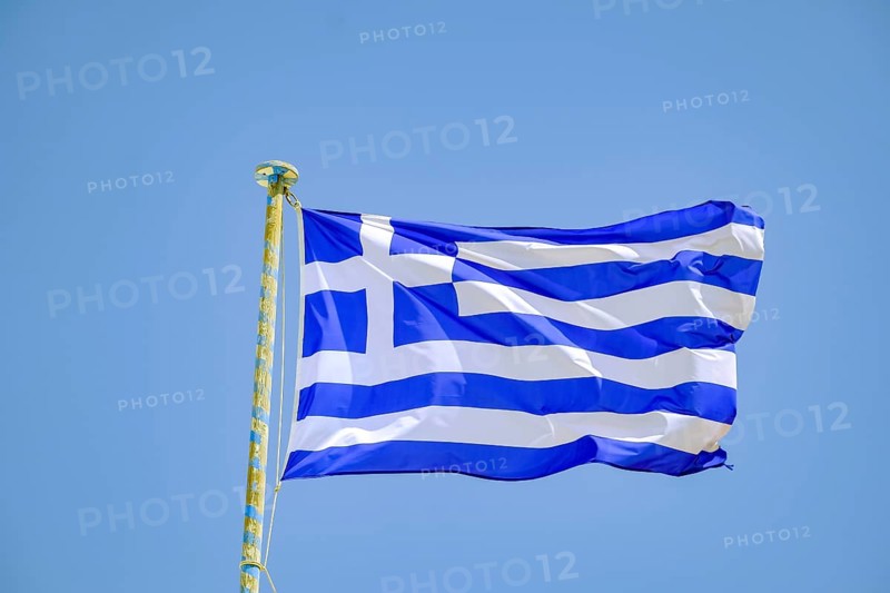 Ano Mera, Mykonos, Cyclades, Greece - Greek national flag fluttering on the flag pole on the roof of Panagia Tourliana monastery forms the center of Ano Mera. The men's convent from the 16th century on the village square in the interior of the island is now a tourist attraction much visited by tourists