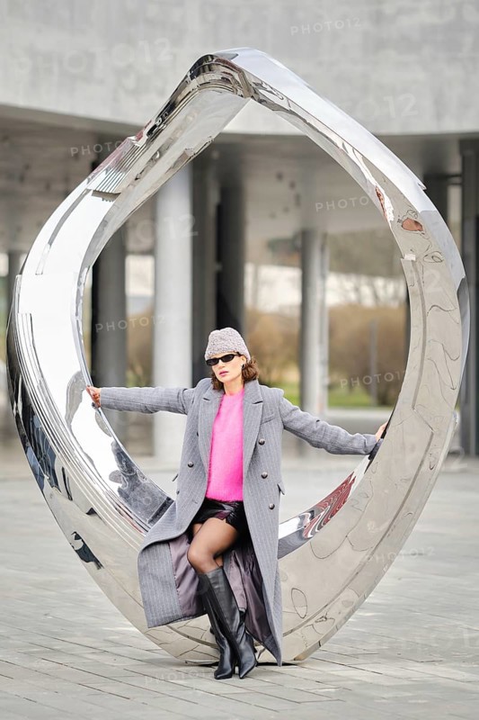 A stylish woman poses sitting on metallic sculpture in a city, showcasing urban street fashion