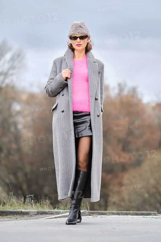 A stylish woman walks down a city street, wearing a pink sweater, unbuttoned long coat, and leather boots. The background features trees and cloudy skies, creating a dynamic urban atmosphere