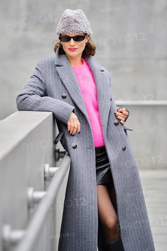 A woman stands confidently on an urban street, dressed in a long gray coat over a vibrant pink sweater. She pairs her outfit with stylish sunglasses and a cozy beanie hat, showcasing modern fashion