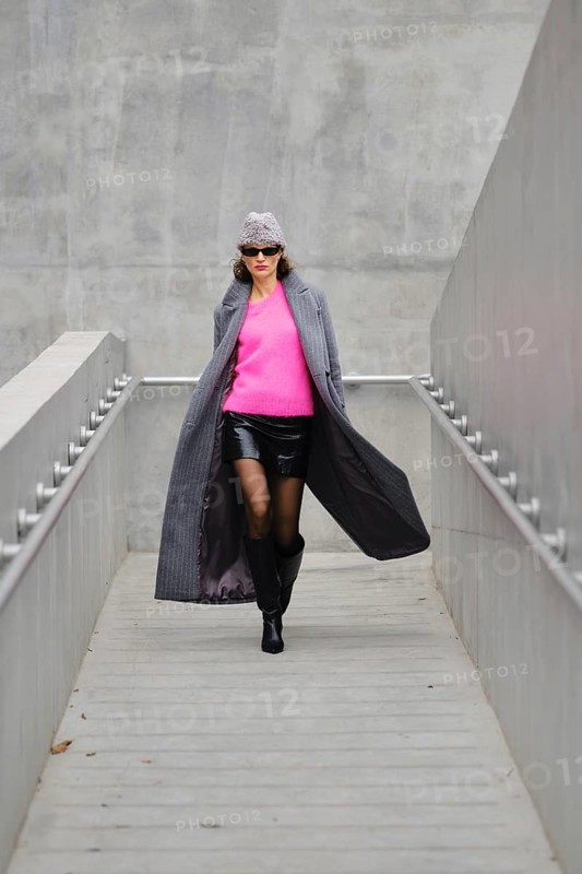 A fashionable woman in sunglasses rushing down a sleek grey walkway, wearing a mini leather skirt, boots, pink sweater and a hat. Her unbuttoned long coat flutters in the wind