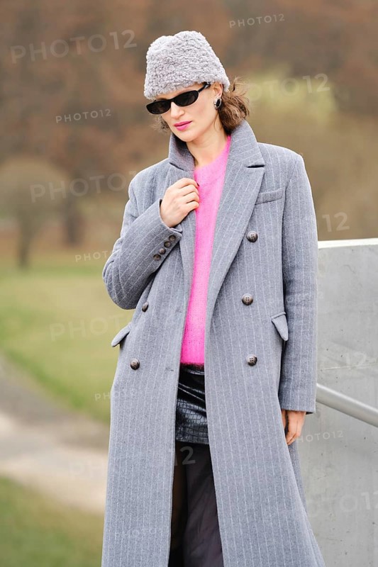 A stylish woman in a long gray coat and bright pink sweater stands outdoors. She wear sunglasses and triangular fur beanie hat while posing against autumn trees and grass