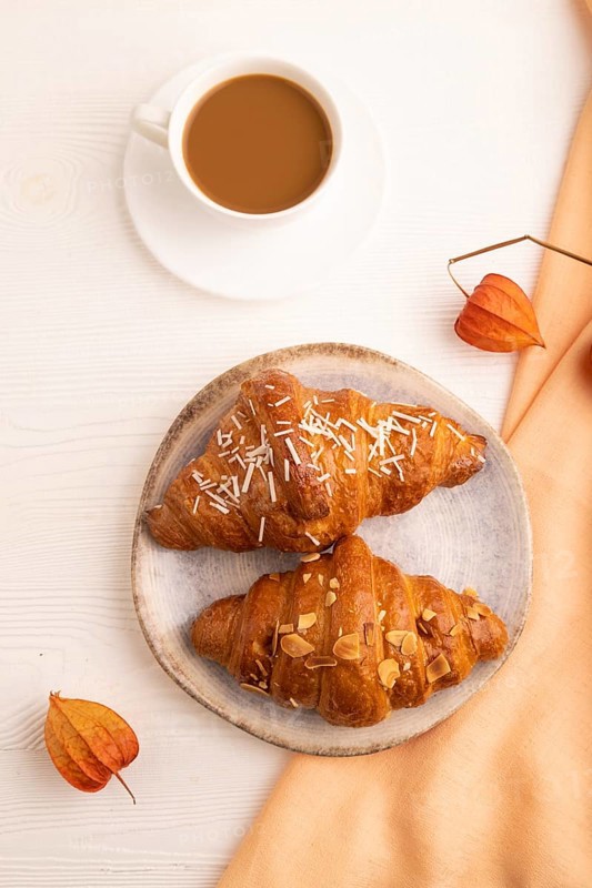 Croissant on blue plate on white wooden background and orange linen textile, cup of coffee, top view, flat lay, close up