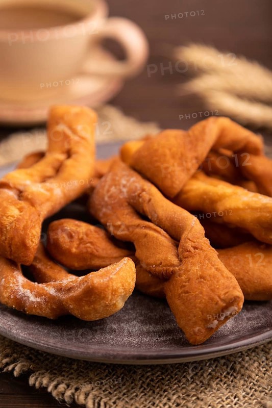 Crunchy biscuit Brushwood cookies sprinkled with powdered sugar on brown wooden background and linen textile, cup of coffee, side view, close up, selective focus