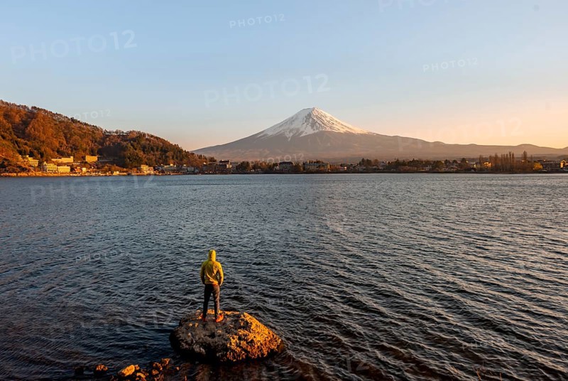 Young man standing on rocks in Lake Kawaguchi, view of Mount Fuji volcano at sunset, Yamanashi Prefecture, Japan