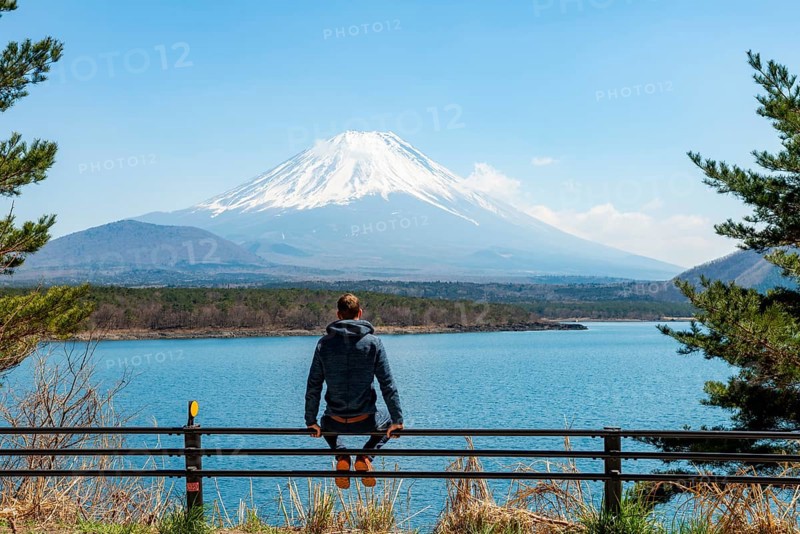 Young man sitting on railings next to a road and looking across the lake to Mt Fuji volcano, Motosu Lake, Yamanashi Prefecture, Japan